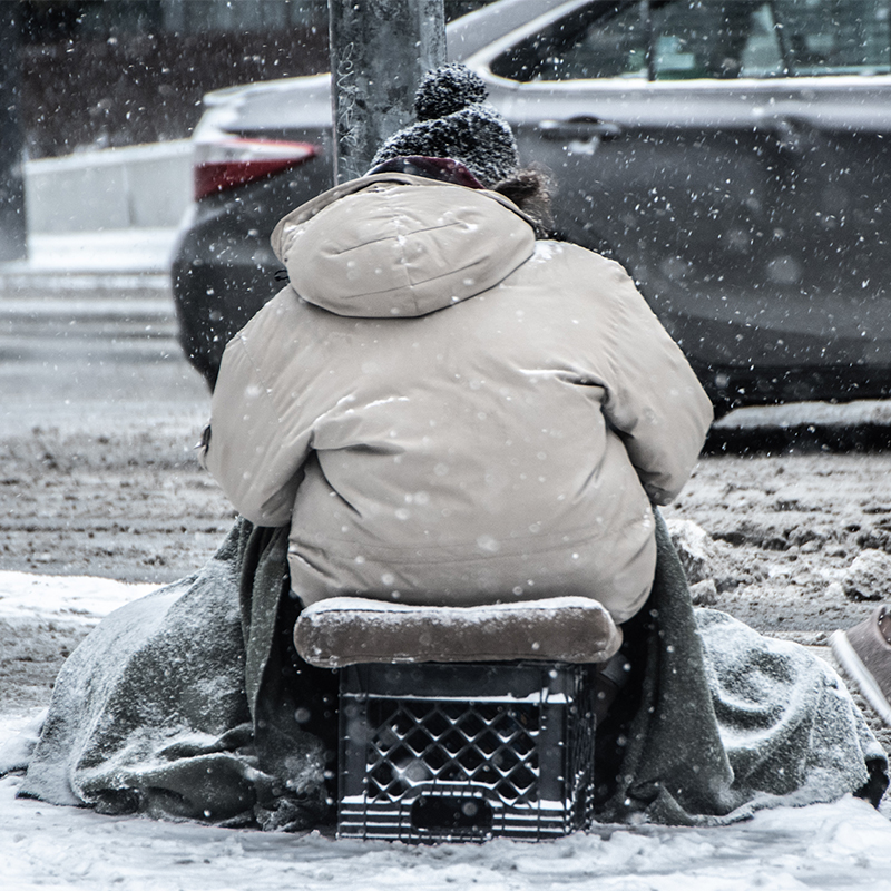 A person sitting on sidewalk during winter weather as another person walks by.