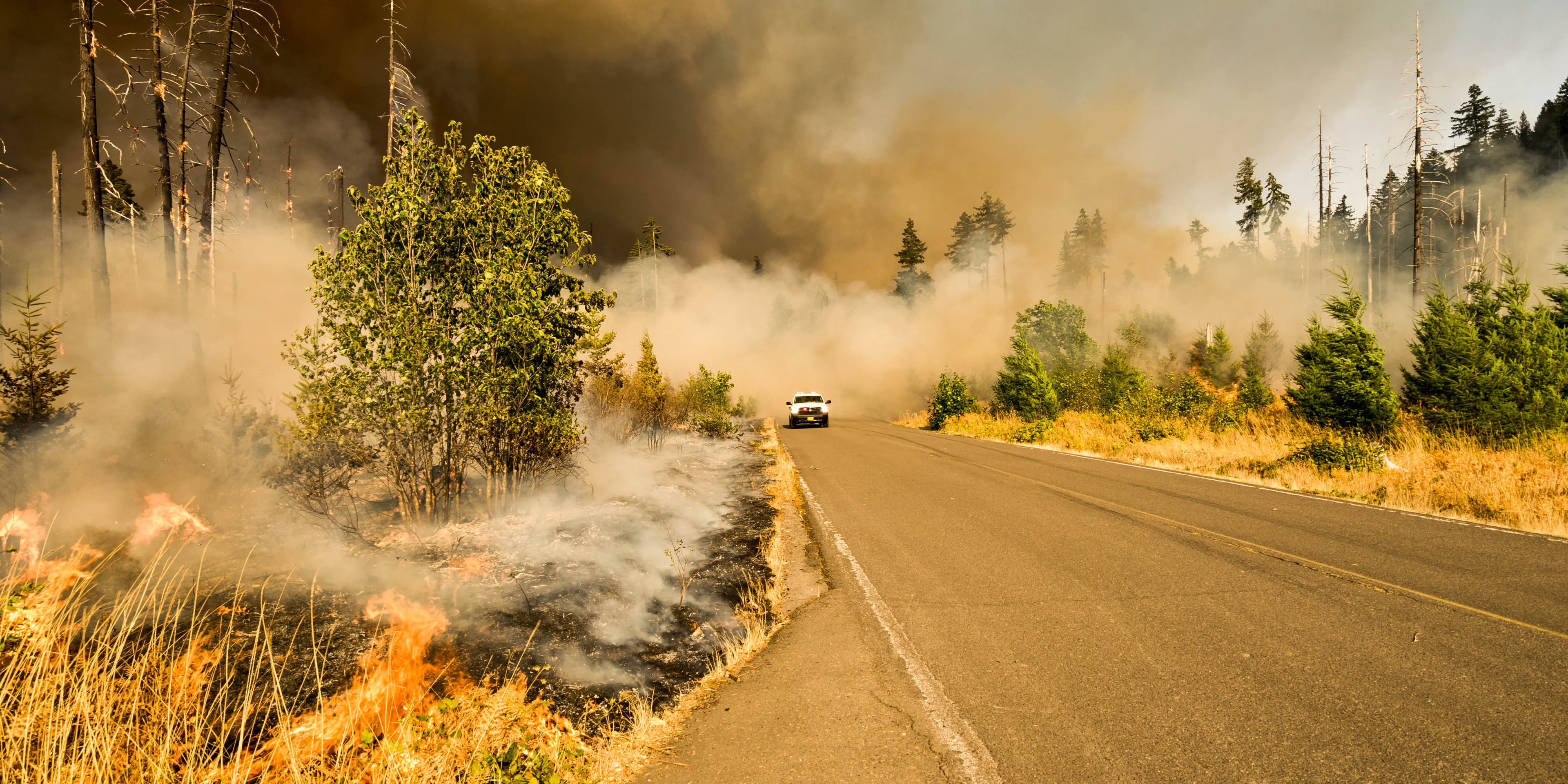 A truck driving on a road surrounded by a wildfire and smoke.